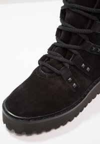 Botte en suède noire avec bout rond, tige texturée, lacets noirs proéminents, clous noirs mats le long des côtés et semelle en caoutchouc robuste.