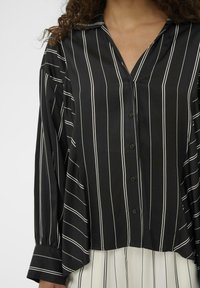 Ženska oblečena v črno srajco na gumbe z belimi navpičnimi črtami ter smetanasto krilo z črnimi navpičnimi črtami.