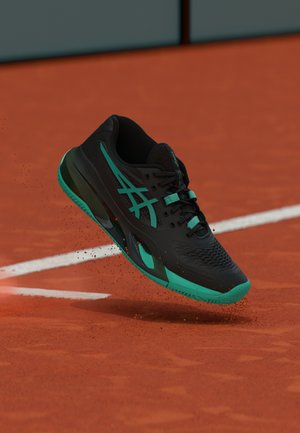 Zwart en teal atletisch schoen zwevend boven een kleiveldtennisbaan met witte begrenzingslijnen, waarbij oranje klei stof opwaait.