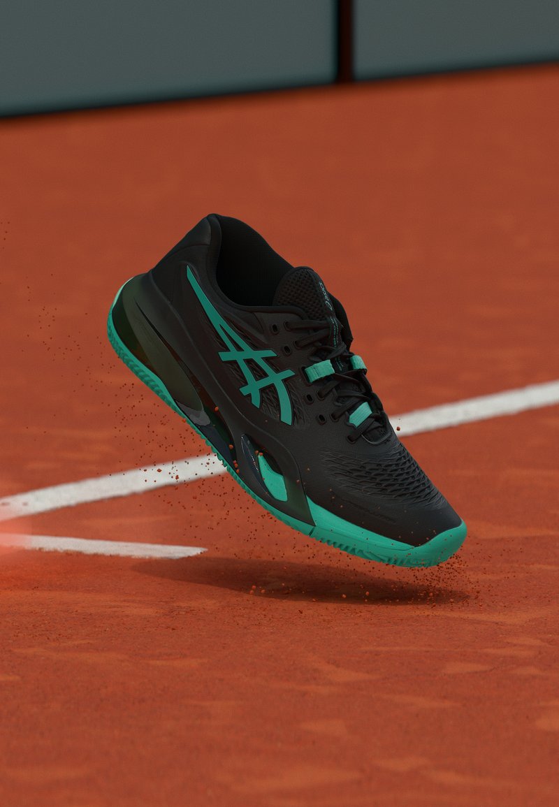 Μαύρο και πετρόλ αθλητικό παπούτσι στον αέρα πάνω από γήπεδο τένις με κοκκινωπό χώμα και λευκές γραμμές οριοθέτησης, σηκώνοντας σκόνη από το πορτοκαλί χώμα.