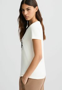 Witte T-shirt met korte mouwen en een ronde halslijn, met een zwart decoratief patroon. Zachte stof, relaxte pasvorm, gedragen met bruine broek.