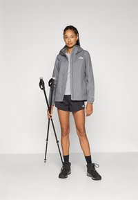 Grijze waterdichte jas met capuchon, zwarte shorts en zwarte wandelschoenen. Houdt trekkingstokken vast, staande tegen een neutrale achtergrond.