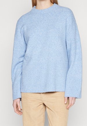 Pull tricoté bleu clair à manches longues porté avec un pantalon beige en velours côtelé sur une personne devant un fond uni.