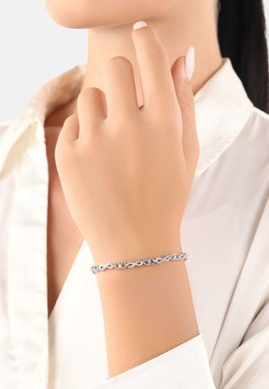 Bracciale in argento con forme a cuore intrecciate e finitura lucida, indossato su una mano di pelle neutra, su uno sfondo chiaro.