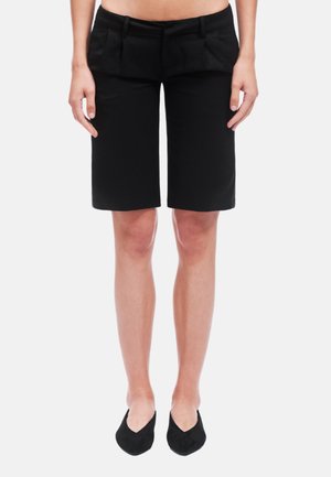 Donna che indossa pantaloncini neri su misura fino al ginocchio e ballerine nere appuntite, in piedi davanti a uno sfondo bianco.