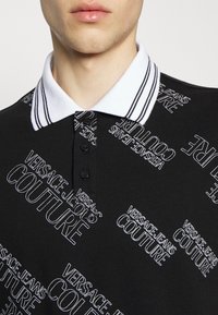 Gros plan d'une personne portant un polo noir à col blanc avec un motif répétitif du texte blanc "Versace Jeans Couture".