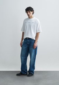 Camiseta blanca de algodón oversize con texto, combinada con jeans azules. La modelo lleva zapatos negros sin cordones. Diseño minimalista sobre un fondo claro.