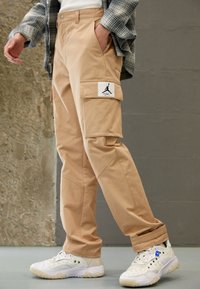 Beiga cargobyxor med en lapp som har en svart logotyp, sidofickor och elastiska muddar, kombinerat med vita sneakers med blå detaljer.