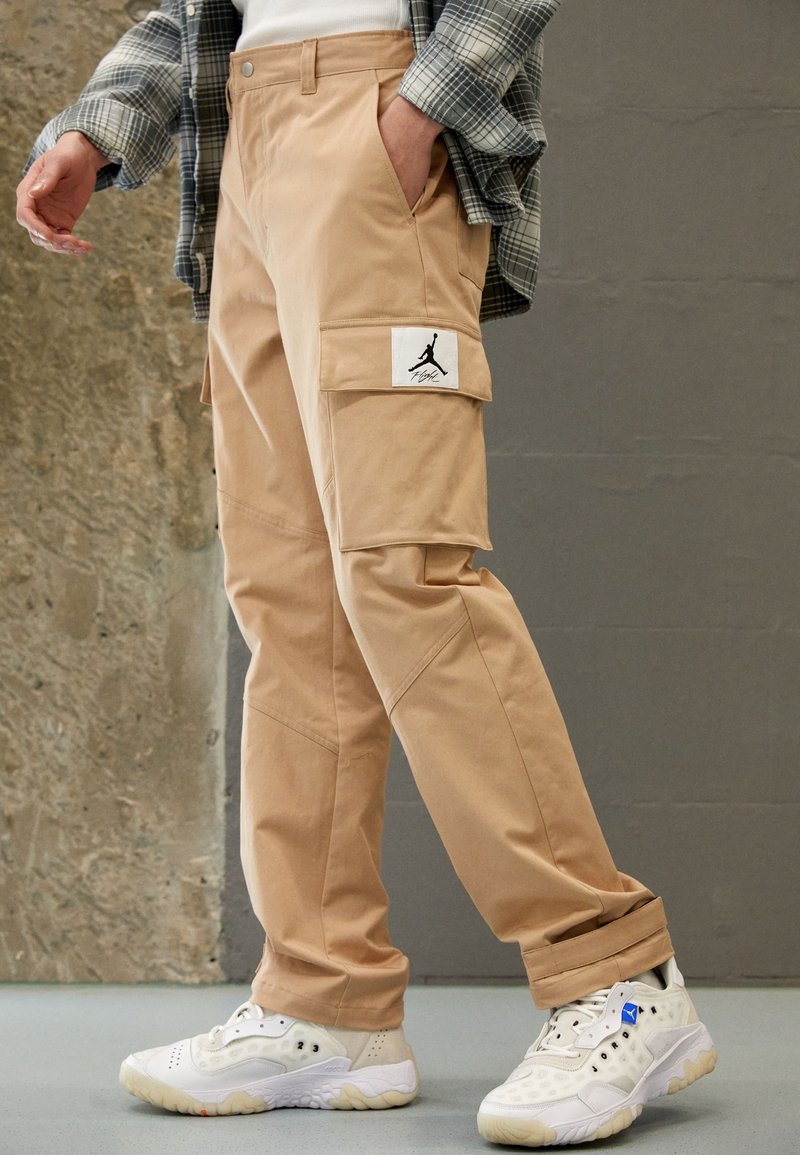 Beiga cargobyxor med en lapp som har en svart logotyp, sidofickor och elastiska muddar, kombinerat med vita sneakers med blå detaljer.