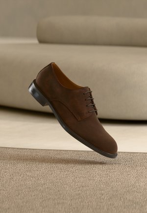 Chaussure habillée en cuir marron à lacets flottant au-dessus d'un tapis beige avec un canapé beige flou en arrière-plan.