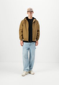 Bruine jas met capuchon over een zwart T-shirt, gecombineerd met lichtblauwe spijkerbroeken en beige sneakers. Bruine pet met logo accentueert de outfit.