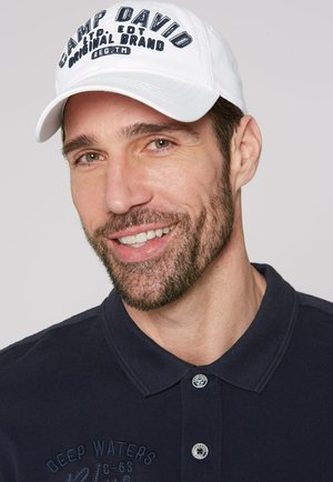 White baseball cap with "CAMP DAVID" logo in dark text. Model wears a navy polo shirt with embroidered pattern and two buttons at the collar.