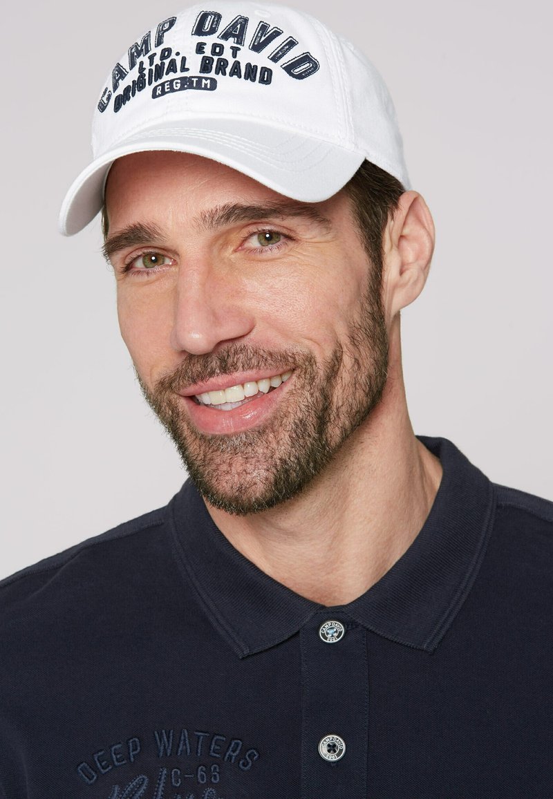 Weißer Baseballcap mit "CAMP DAVID"-Logo in dunkler Schrift. Das Modell trägt ein navy-blaues Poloshirt mit gesticktem Muster und zwei Knöpfen am Kragen.