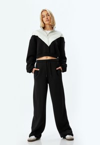 Sudadera corta negra con diseño en forma de chevron gris, cuello con media cremallera, combinada con pantalones de chándal sueltos negros, que cuentan con bolsillos laterales y cordón.