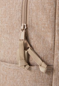 Mochila de tela beige con superficie texturada, que presenta un cierre y detalles de correa en tono marrón. El diseño incluye un bolsillo lateral con herrajes ajustables.
