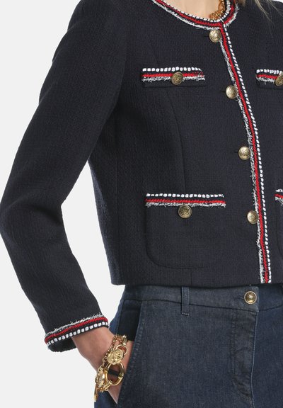 Veste marine texturée avec boutons dorés, bordures blanches et rouges, et poches à rabat. Caractérisée par un design court et des détails complexes.