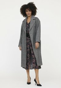 Langer grauer Fischgrätmantel mit übergroßen Revers, getragen über einem Blumenkleid. Verfügt über weite Ärmel und eine lockere Passform, ergänzt durch schwarze High Heels.