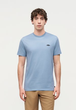 Mladý muž s kučeravými tmavými vlasmi, oblečený v svetlomodré tričko s krátkym rukávom a béžové nohavice, stojaci pred jednoduchým pozadím.