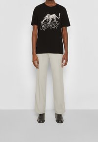 Mężczyzna noszący czarną koszulkę z białym nadrukiem warczącej pantery i napisem "LOVE NOW", beżowe spodnie i czarne buty.