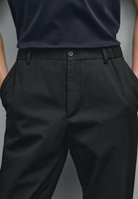 Black trousers made of smooth fabric featuring a button front, belt loops, and side pockets, with a relaxed fit design.