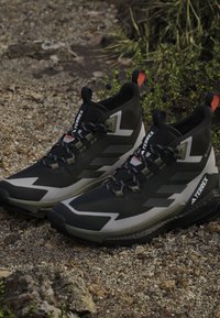 Μαύρα και γκρι παπούτσια πεζοπορίας Adidas Terrex με κορδόνια σε βραχώδες, χαλίκι έδαφος με πράσινα φυτά στο φόντο.