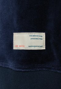 Marineblauwe stof met een zachte textuur. Bevat een genaaid label in crème met rode en muntgroene tekst met de opschriften "odd molly," "celebrating yourselves," en "uncorporated."