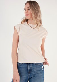Top corto a manica corta beige chiaro in morbido cotone, con una vestibilità comoda e bordi delle maniche leggermente arrotolati, abbinato a jeans blu strappati.