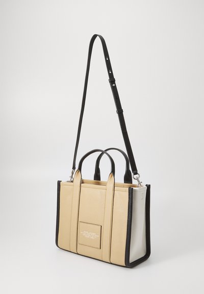 Beige leather handbag with black accents; features two top handles, detachable strap, and logo patch on the front. Structured design.