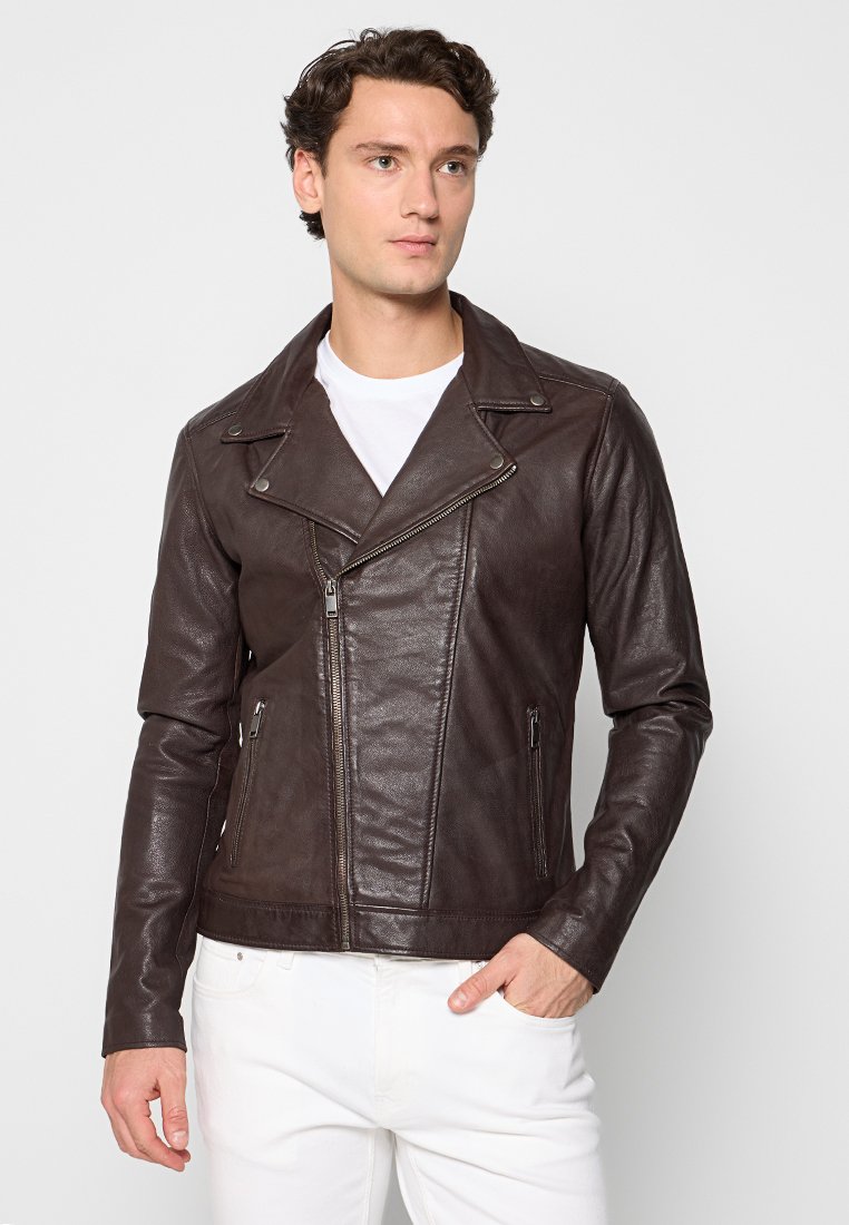 Man met donker krullend haar, gekleed in een donkerbruine leren motorjack over een wit shirt en witte broek, staande met één hand in de zak.