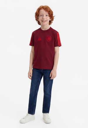Rødhåret dreng iført en mørkerød Arsenal T-shirt, blå jeans og hvide sneakers, der står og smiler mod en hvid baggrund.