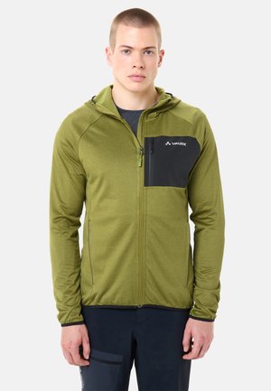 Giovane uomo che indossa una felpa con cappuccio verde oliva con cerniera e una tasca nera sul petto, in piedi di fronte a uno sfondo semplice.