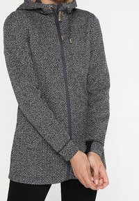 Strikjakke i grå og sort med hætte, fuld lynlås og elastiske manchetter. Har et struktureret mønster og trædetaljer på lynlåsen.