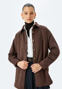Chaqueta de ante marrón con un diseño de cuello, adornada con cinco botones dorados en la parte delantera. Combinada con un cuello alto marrón acanalado debajo.