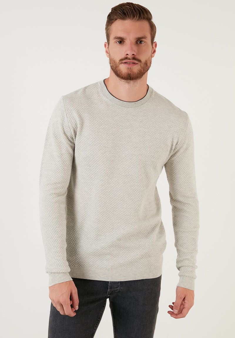 Ljusgrå texturerad stickad tröja med rund halsringning och långa ärmar. Har ribbade manschetter och fåll, kombinerad med mörka jeans.