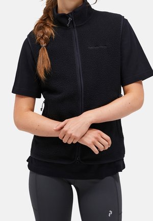 Kobieta z warkoczami, ubrana w czarną kamizelkę polarową na zamek, na czarnej koszulce z krótkim rękawem i ciemnoszare legginsy, z rękami luźno skrzyżowanymi.