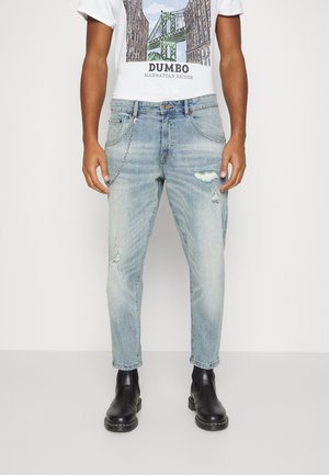 Homme portant un jean bleu clair déchiré avec un accessoire chaîne, des bottes en cuir noir et un t-shirt blanc avec un motif du pont de Manhattan.