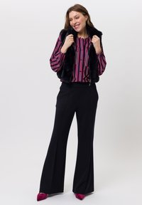 Zwarte nep-bontvest gedragen over een rood-blauw gestreept blouse, gecombineerd met zwarte wijde pantalon en bordeauxrode pumps met spitse neus.