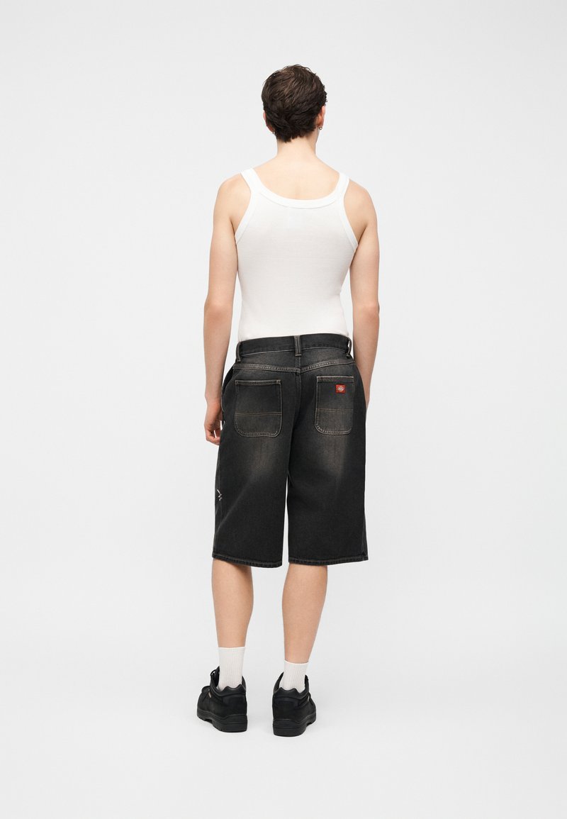 Person trägt ein weißes Tanktop, schwarze lange Jeansshorts, weiße Socken und schwarze Sneakers, steht mit dem Rücken zu einem schlichten Hintergrund.
