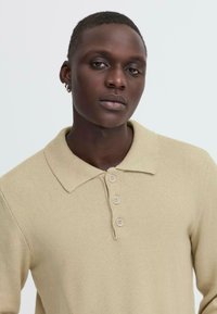 Tricou polo tricotat de culoare bej, cu guler, având un plastron cu trei nasturi și țesătură texturată. Nasturii sunt de culoare deschisă și rotunzi.