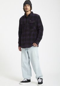 Camisa de franela a cuadros negra y morada con dos bolsillos en el pecho, combinada con vaqueros holgados de color azul claro y zapatillas negras. Tejido texturizado.