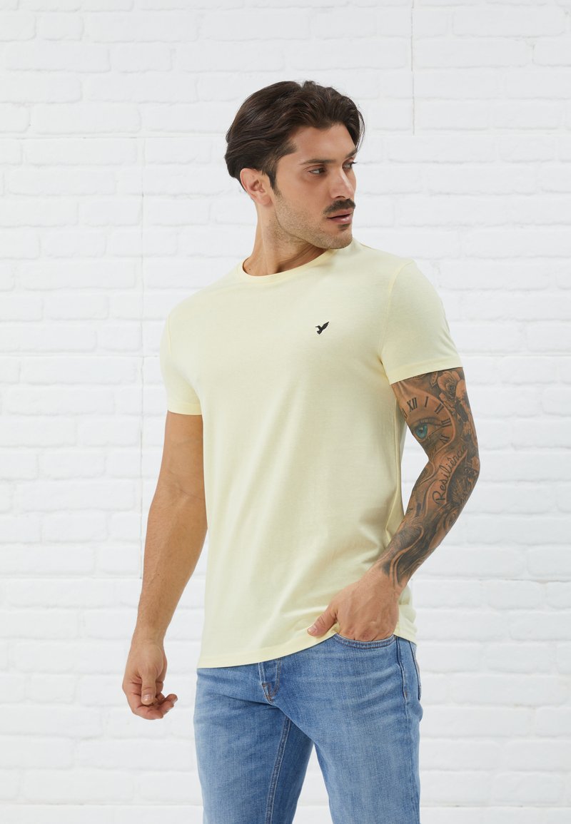 Light yellow short-sleeve t-shirt with a round neckline and a small black logo on the left chest. Smooth fabric, worn with blue jeans.