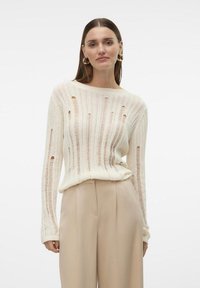 Pull en tricot crème avec des rayures verticales et des détails découpés ; associé à un pantalon beige clair, affichant une coupe décontractée.