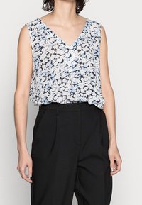 Donna che indossa una blusa floreale senza maniche con scollo a V, caratterizzata da fiori blu chiaro e bianchi, infilata in pantaloni neri con vita alta.