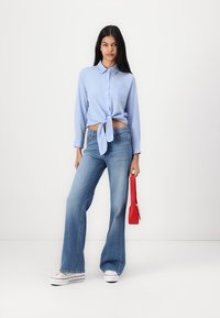 Lys blå skjorte med knapp som er knyttet rundt livet, kombinert med vide blå jeans og hvite sneakers. Holder en liten rød håndveske.