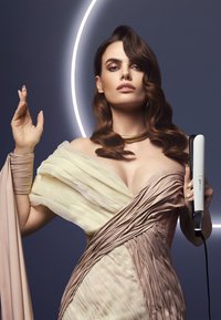 Piastra per capelli in mano, modello indossa un abito drappeggiato con tessuti beige e crema. Lo sfondo presenta una luce circolare soffusa e luminosa.