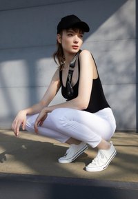 Black sleeveless bodysuit, white fitted ankle pants, and white sneakers with black stripes. Model squats on a concrete surface, wearing a matching cap.