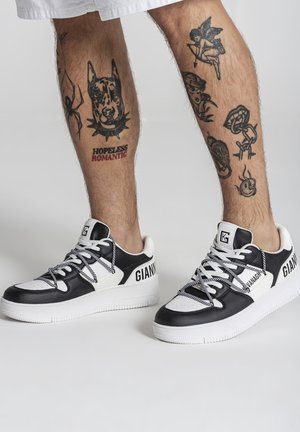 Beentattoo's met zwarte en witte Gianni-sneakers, met daarop tatoeages van een hond, spin, Cupido, lippen en de tekst "Hopeless Romantic".