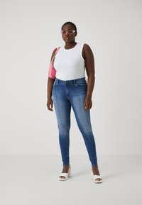 White sleeveless top, blue skinny jeans, and white heeled sandals. Pink handbag with silver hardware. Clear rectangular sunglasses.