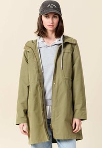 Olivgrön huvjacka med dragkedja fram, sidofickor och guldfärgade detaljer, lager-på-lager över en ljusgrå sweatshirt och ljusblåjeans.