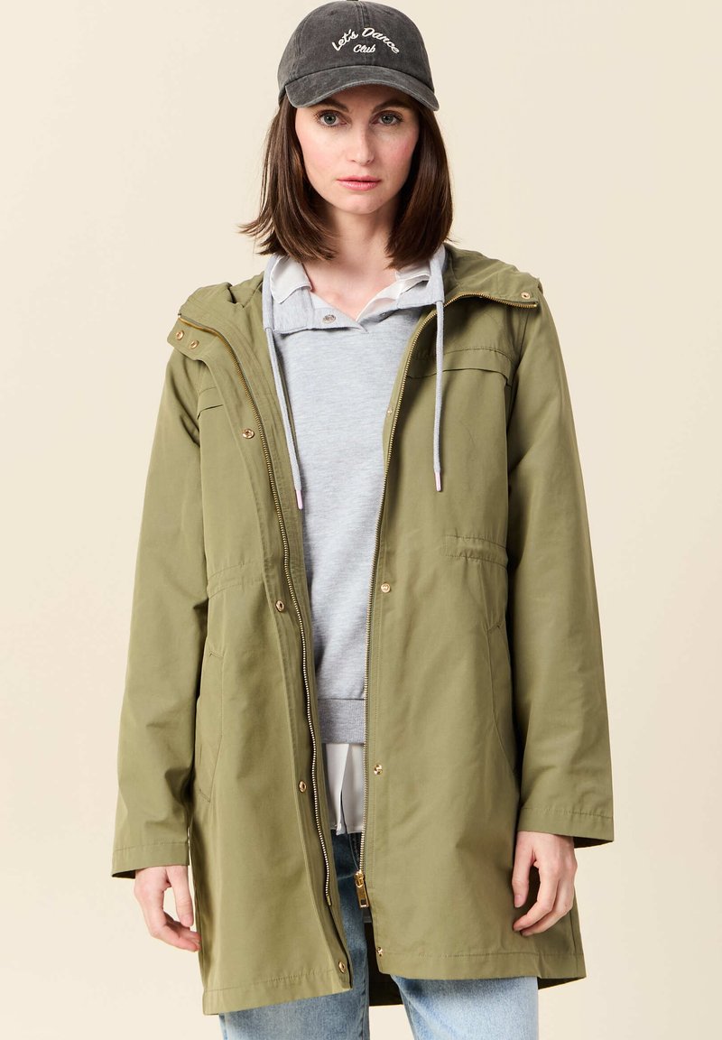 Olivgrön huvjacka med dragkedja fram, sidofickor och guldfärgade detaljer, lager-på-lager över en ljusgrå sweatshirt och ljusblåjeans.